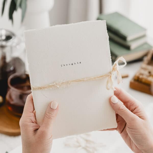 an off white journal with a deckled edge, a silk ribbon tied in a bow to close it and the words 'thoughts' typed on the cover - By Blue Stiggy