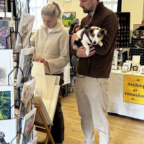 A man and woman look at a display of prints. The man is holding a small dog.