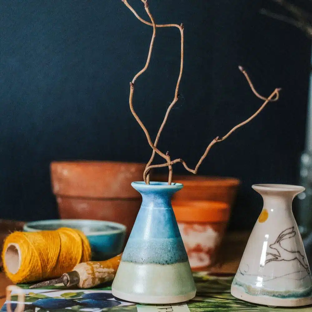A photo of a bud vase with twigs in it sitting on a gardening book with plant pots and gardening string in the back ground. The vase is decorated in blue and turquoise glazes.