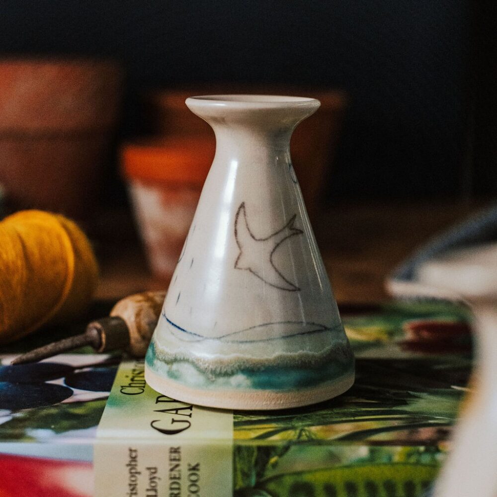 A bud vase decorated with an off white glaze and a swift bird design. Sitting on a gardening book with plant pots and string in the background.