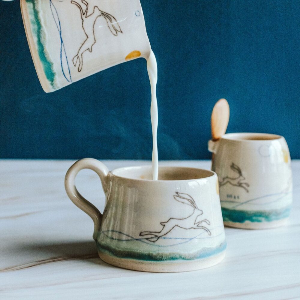 A collection of ceramics decorated with running hares. the handmade cup is having milk poured into from a milk jug. There is a sugar bowl in view behind the cup.