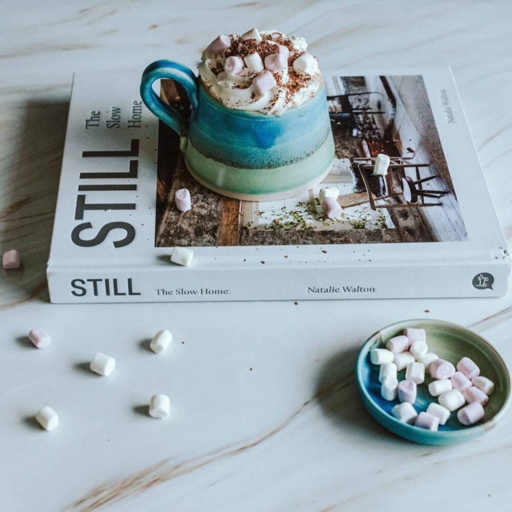 A small bowl full of mini marshmallows next to a handmade mug full to the brim with hot chocolate with cream and marshmallows on top.