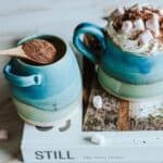 A pot full of chocolate flakes next to a handmade mug full to the brim with hot chocolate with cream and marshmallows on top.