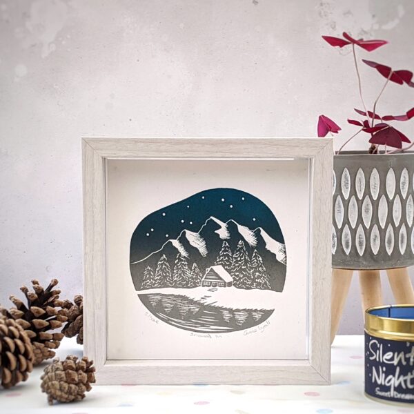 Rose and Hen, a 6 x 6 inch linocut print of a log cabin by a lake with woods and snow capped mountains in the background and stars in the sky.