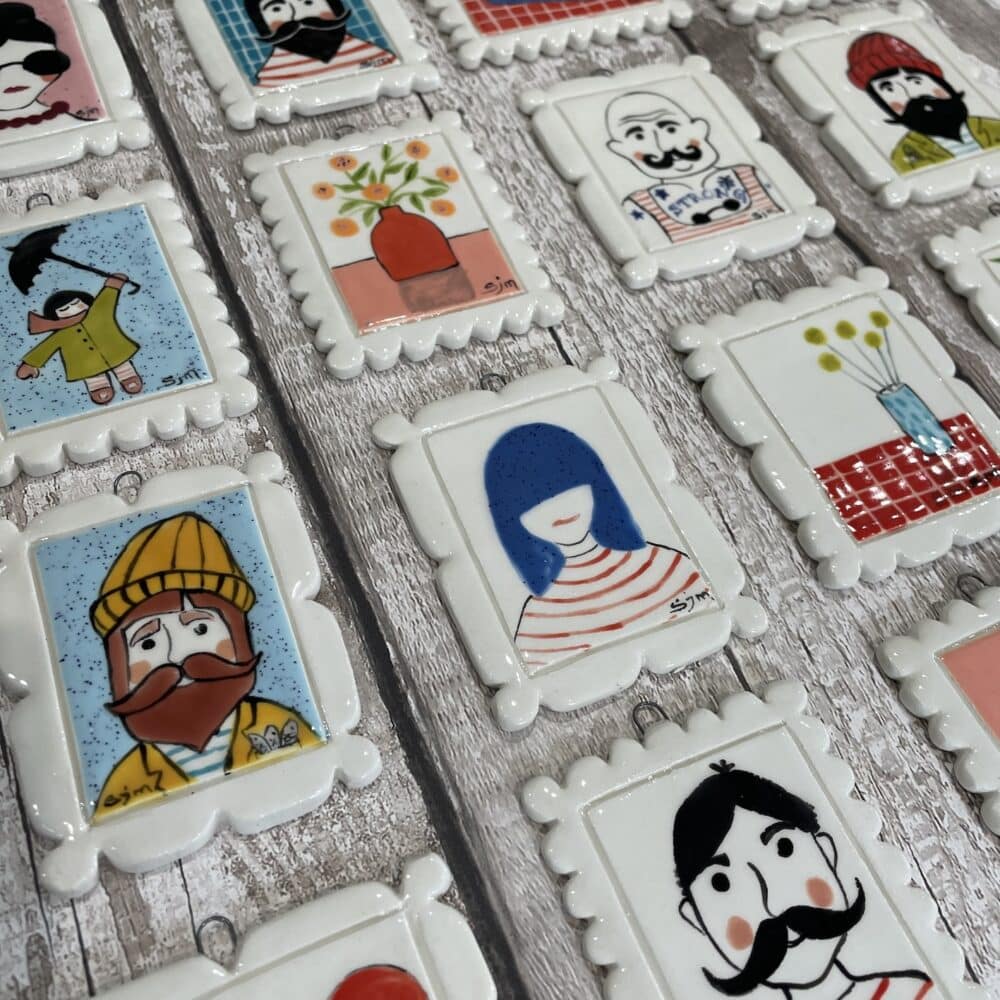 A collection of small rectangular ceramic mini picture frame with hand-painted illustrations, laid out on a rustic wooden surface. The Mini Masterpieces have a glossy glaze finish with decorative, scalloped edges and feature various whimsical designs, including stylized portraits of people with striped shirts, beards, and mustaches, as well as objects like flowers in vases, and sandals. The artwork has a playful, folk-art aesthetic with bold colors and simple lines