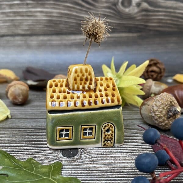 Handmade miniature ceramic green house with a honey coloured door, windows and roof. The house has a chimney that can hold a flower or dried stem, displayed on a rustic wooden background with autumn leaves and acorns