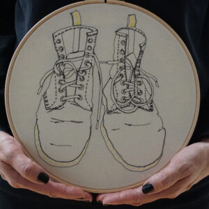 Gemma Rappensberger holding a Dr Martens Boots embroidered illustration in free motion machine embroidery in black thread with yellow hand embroidery details on calico, displayed in a 10" wooden hoop, held by Gemma Rappensberger.
