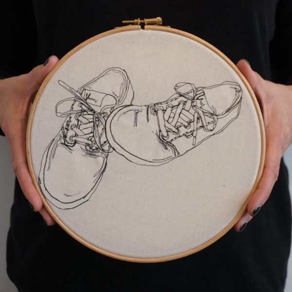 Gemma Rappensberger holding her Vans trainers embroidered illustration using free motion machine embroidery in black thread on calico displayed in a 9" wooden embroidery hoop, being held by Gemma Rappensberger