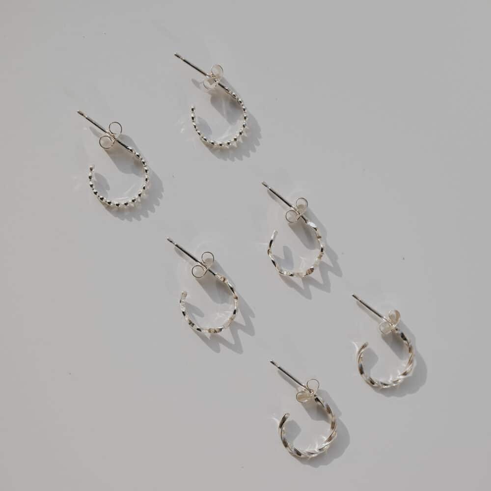 Silver mini hoop earrings flat lay with wavy, dot, and twisted designs arranged diagonally on grey background