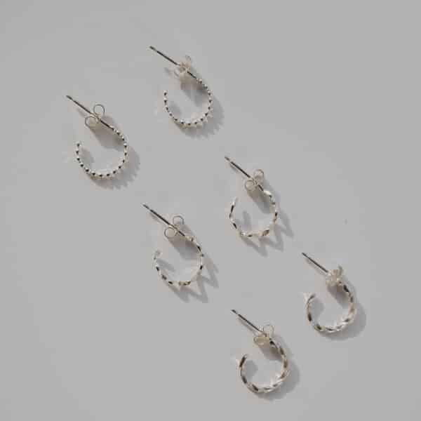 Silver mini hoop earrings flat lay with wavy, dot, and twisted designs arranged diagonally on grey background
