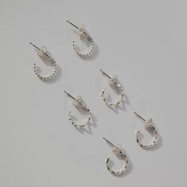 Silver mini hoop earrings flat lay with wavy, dot, and twisted designs arranged diagonally on grey background