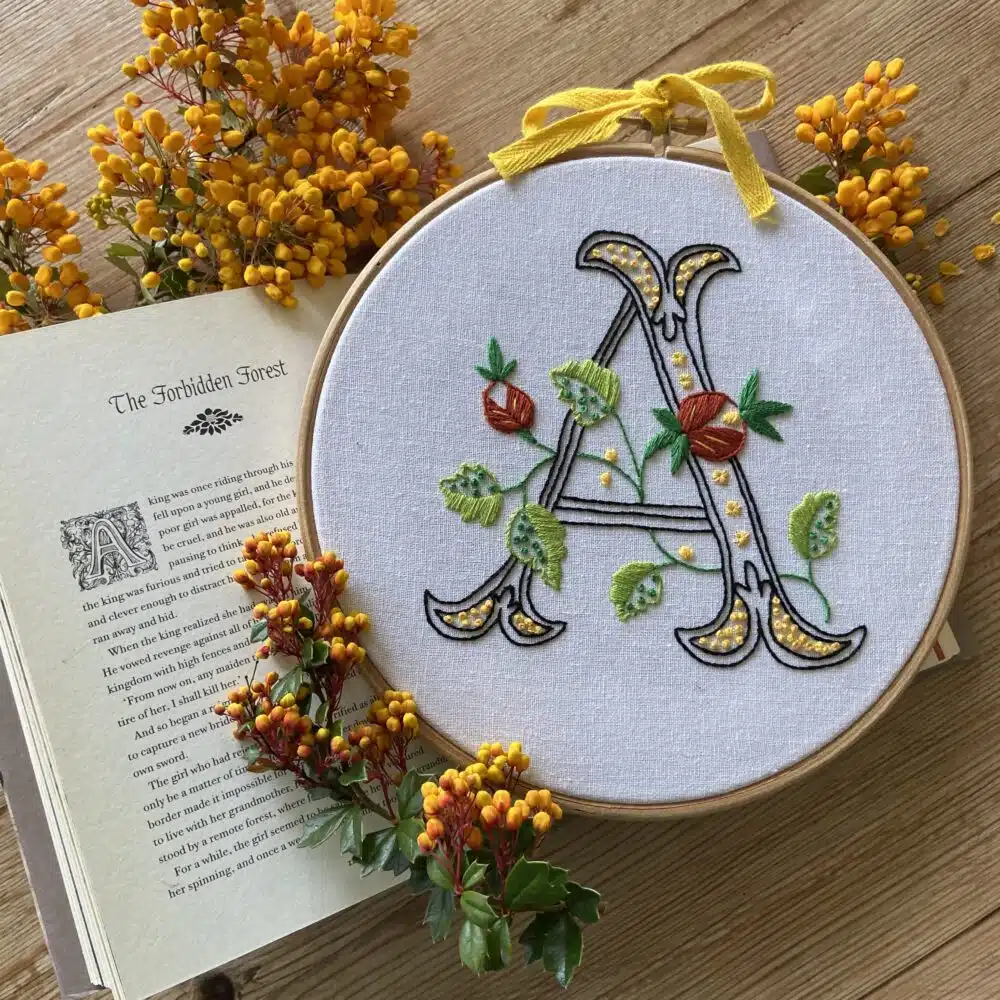 An embroidery kit featuring a decorative embroidered initial, complete with fabric, embroidery threads, needle, hoop, and instructions.