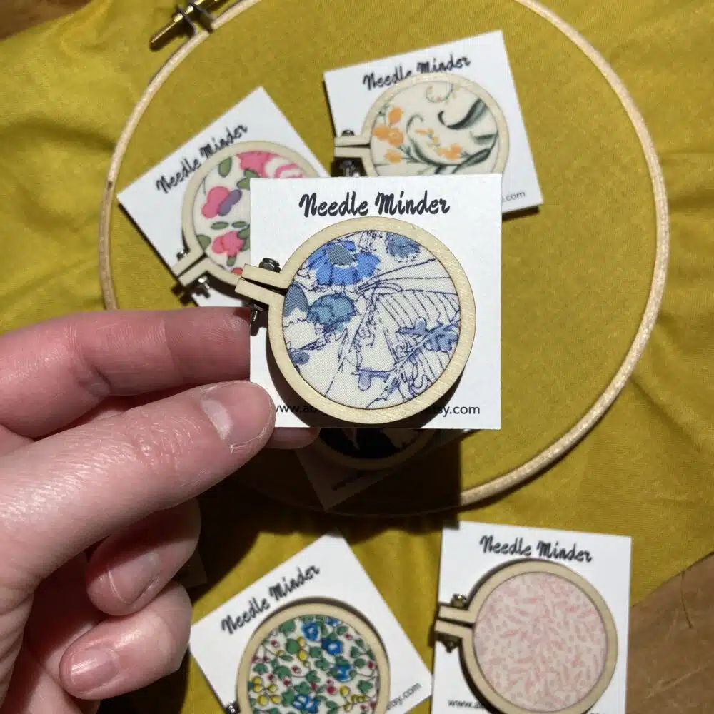 A collection of handmade mini embroidery hoop needle minders covered in colourful Liberty fabrics, designed to hold embroidery needles securely while stitching.