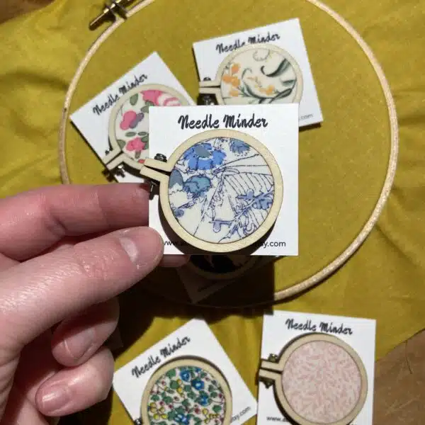A collection of handmade mini embroidery hoop needle minders covered in colourful Liberty fabrics, designed to hold embroidery needles securely while stitching.