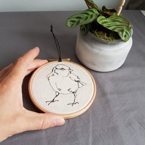 Gemma Rappensberger holding an embroidered illustration in free motion machine embroidery in black cotton with hand embroidery detail on calico in a wooden embroidery hoop