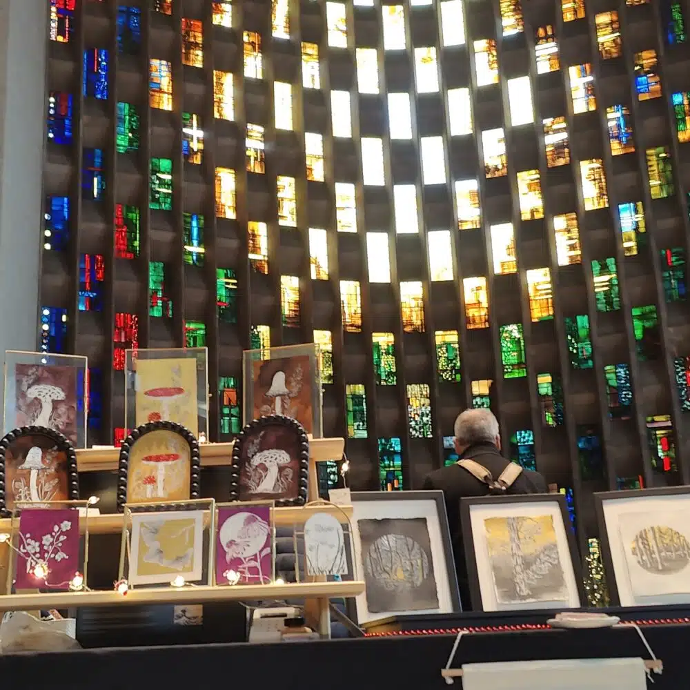 Art stall displaying framed artwork with a mix of different trees, mushrooms and flowers, in front of the Coventry Cathedral stained glass wall
