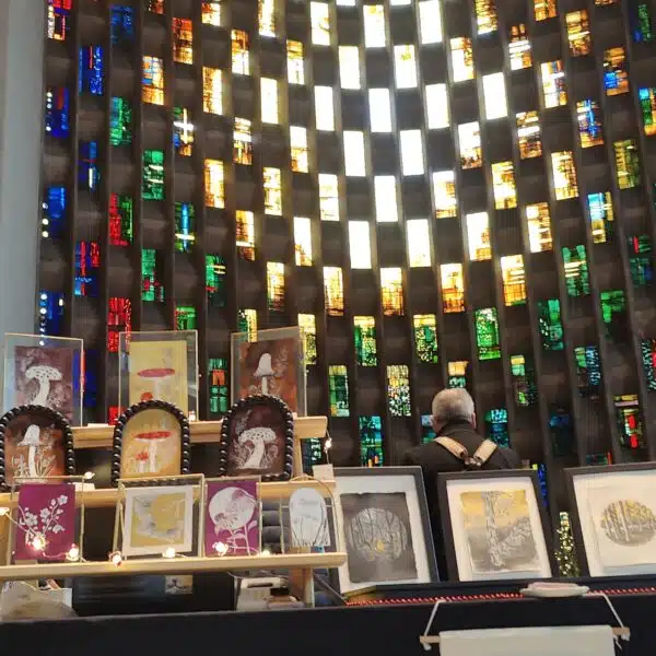 Art stall displaying framed artwork with a mix of different trees, mushrooms and flowers, in front of the Coventry Cathedral stained glass wall