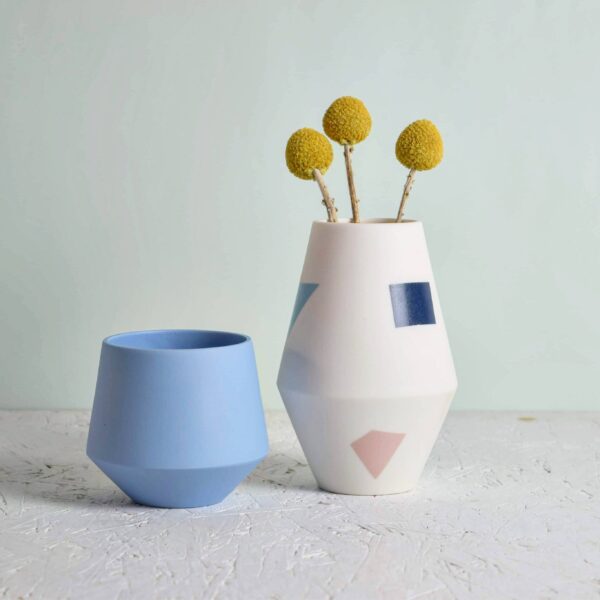 in the photo is a ceramic blue cup and a 20cm high white vase decorated with geometric blocks of baby pink, navy blue, and light blue. in the vase is 3 dried flower stems visualising the objects intended use.