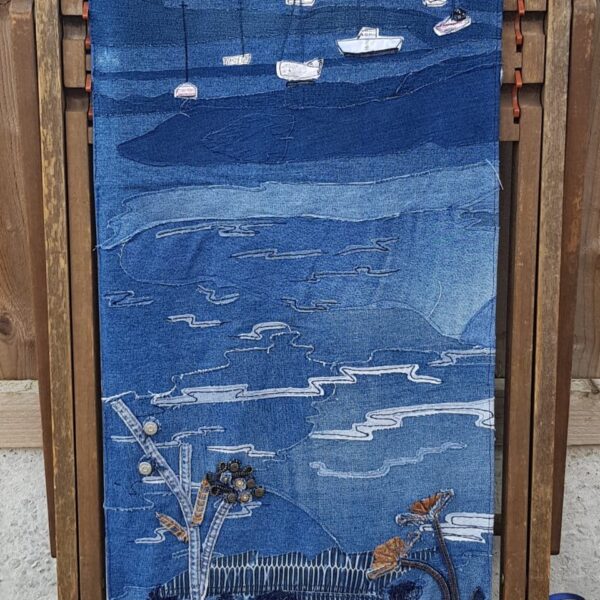 A denim stitched landscape portrait with a riverbank in the foreground, mudflats and boats in the middle and boats on the river on the horizon. Opposite shore lined with trees in the background. A traditional wooden framed folding deck chair