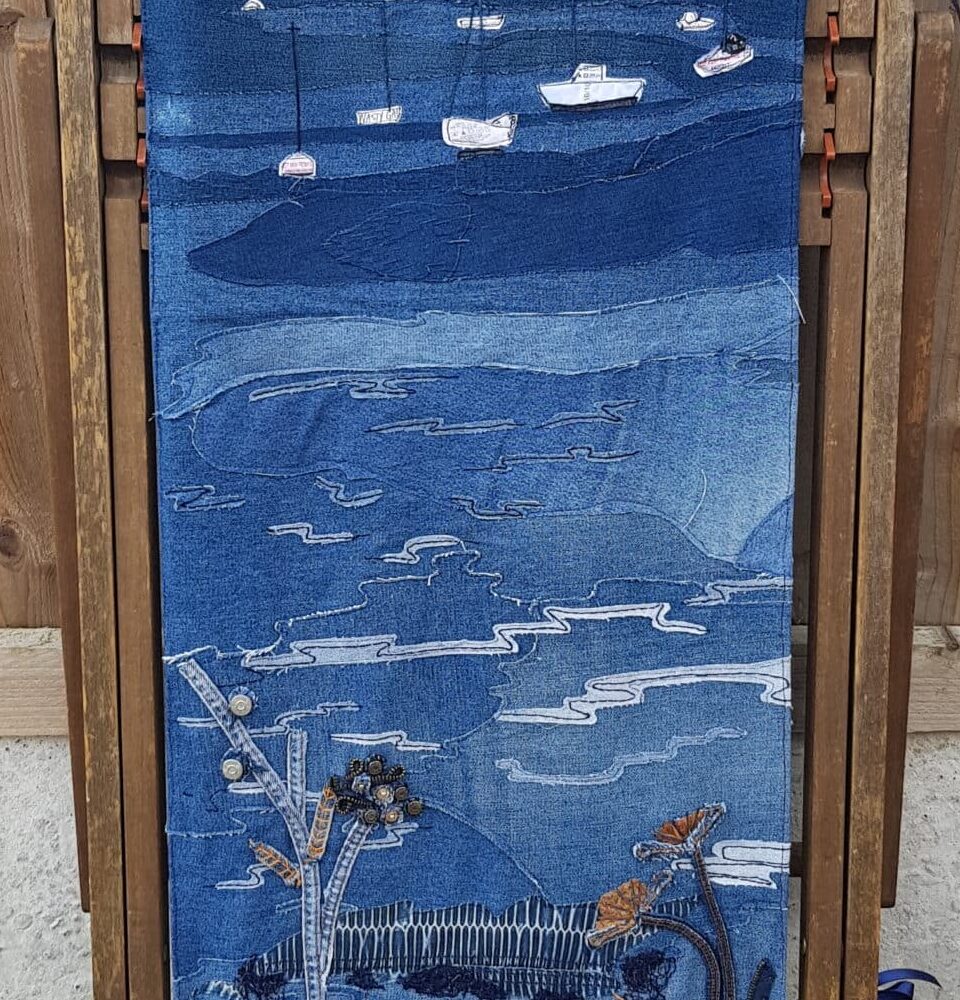 A denim stitched landscape portrait with a riverbank in the foreground, mudflats and boats in the middle and boats on the river on the horizon. Opposite shore lined with trees in the background. A traditional wooden framed folding deck chair