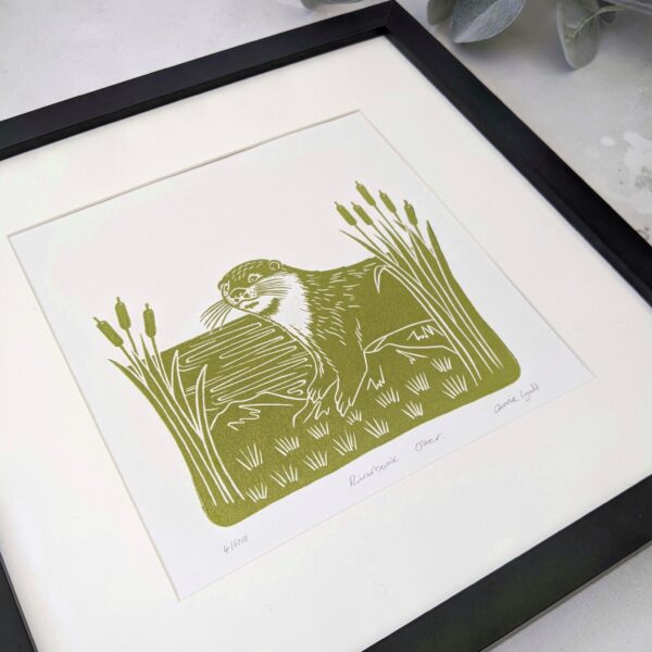A linocut print of an otter on a riverbank framed by bulrushes in green - rose and hen