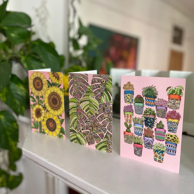 Three card designs, sunflowers, tropical leaves and pot plants, lined up next to each other on fireplace with plant and mirror in background