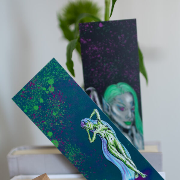 Diva Collection Bookmarks by Hannah Kate Makes. Two bookmarks poking out the top of books on a shelf. One showing the green Drama Queen illustration, the other a green haired dark angel.