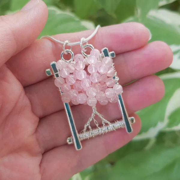 PeachTreePig - Oblong shaped Rose Quartz Tree of Life pendant necklace, with a hallmarked Sterling Silver frame and round bead detailing. Held in hand against a leafy background (© PeachTreePig)