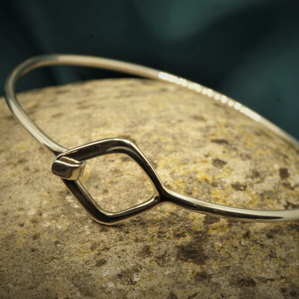 Oval sterling silver bangle with kite shape detail closure resting on a stone