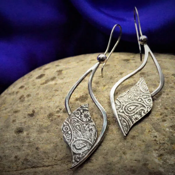 Paisley textured wave shaped silver drop earrings resting on stone