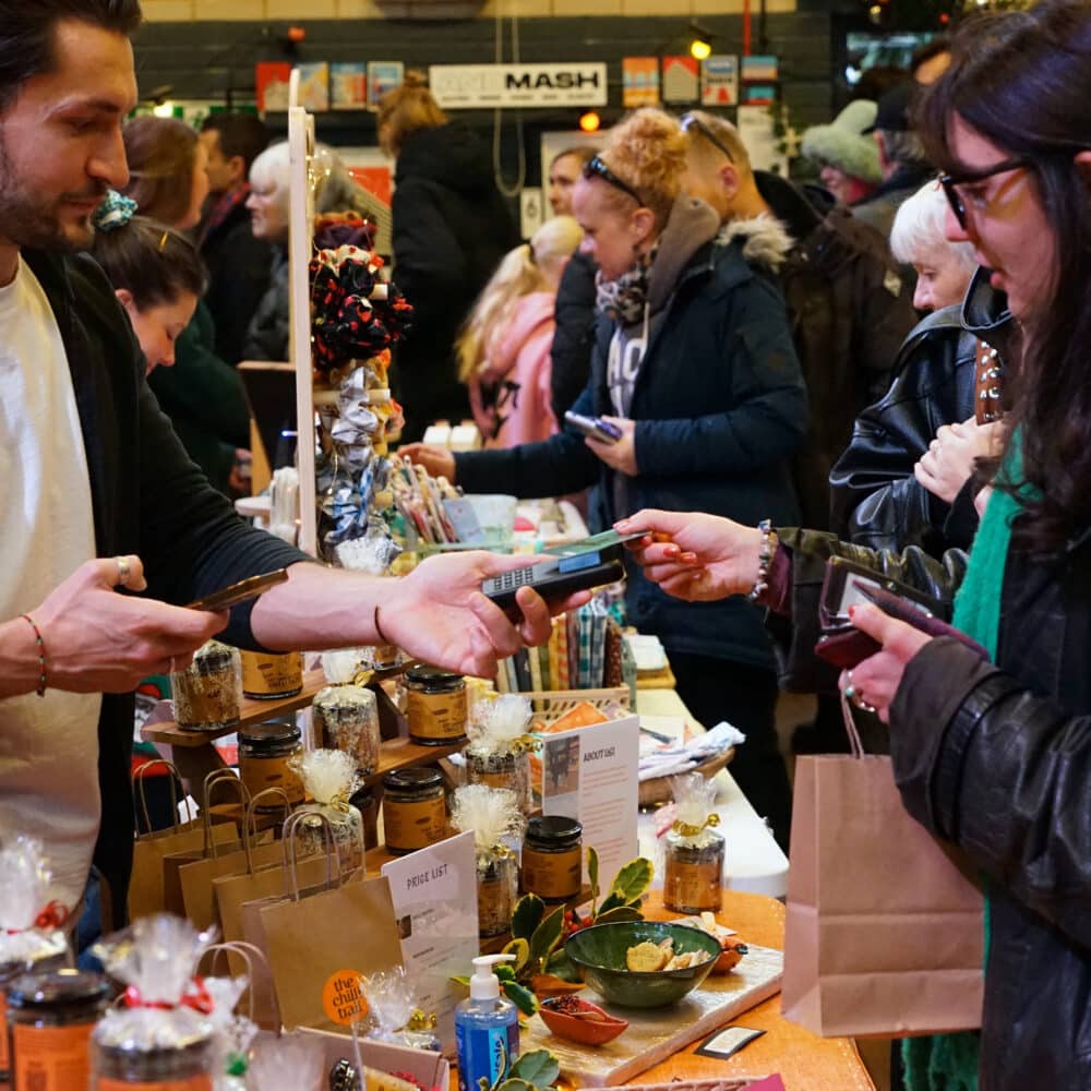 Customer paying an independent food producer at a busy festive makers fair stall inside Wagner Hall in Brighton.