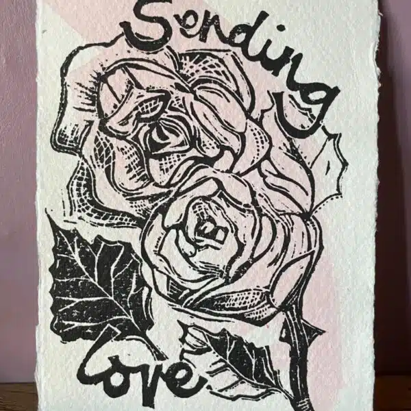 Mandy Benton Printmaker, 'Sending Love' Linocut Print. A small linocut print on a beautiful handmade paper depicting roses and the message 'Sending Love'. Printed in black ink over a sweep of pink watercolour paint.