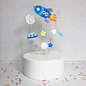 A blue and silver space theme cake topper with rocket ship, planets, stars and alien spaceship. The cake topper is personalised with the birthday age on the rocket and name on a shooting star i