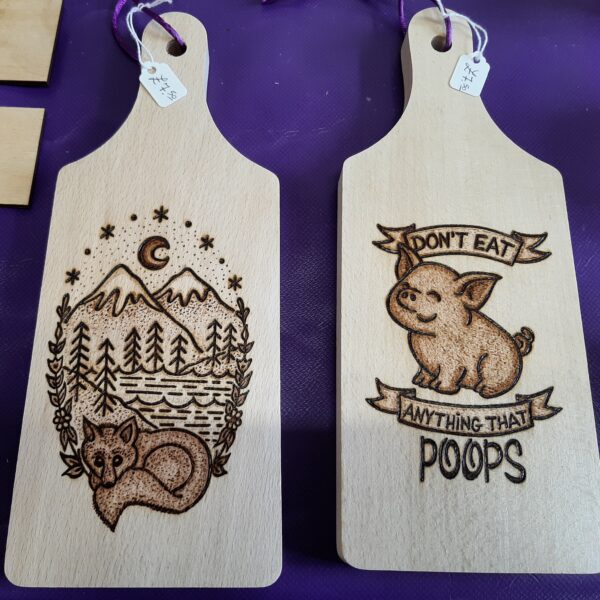 Two pyrography chopping boards with images and a slogan. The first shows a fox curled up beneath a mountain range and the second is of a cheerful cartoon pig with the message "Don't eat anything that poops!" Found at Newark Vegan & Ethical Market