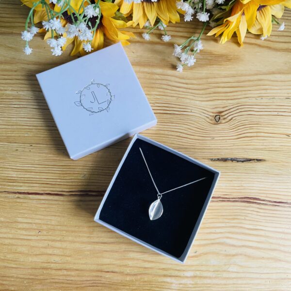 Wooden table top with fake sunflowers, showing a pearlescent ivory branded JL box open with a sterling silver leaf pendant inside