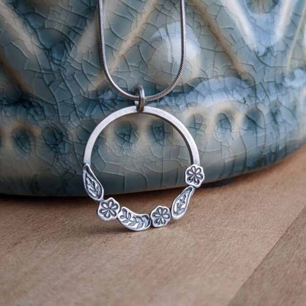 A silver hoop pendant with stamped leaves and flowers, leaning against a blue vase on a wood table.