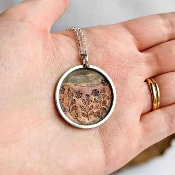 A copper disc pendant, surrounded by a silver hoop, being shown on a hand.