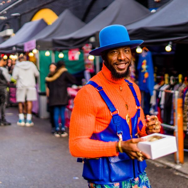 Black man wearing blu hat and blue top over and orange polo neck jumper Black Owned Market with Bohemia Place