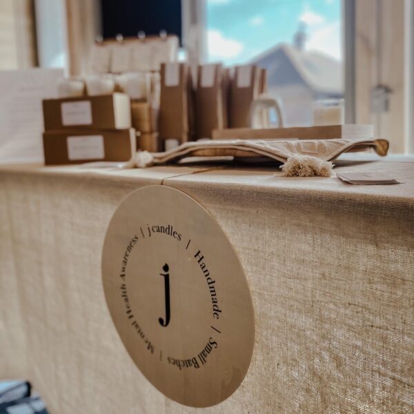 Table covered in a hessian cloth with a wooden plaque showing a lower case j and the words "j candles, handmade, small batches, mental health awareness"