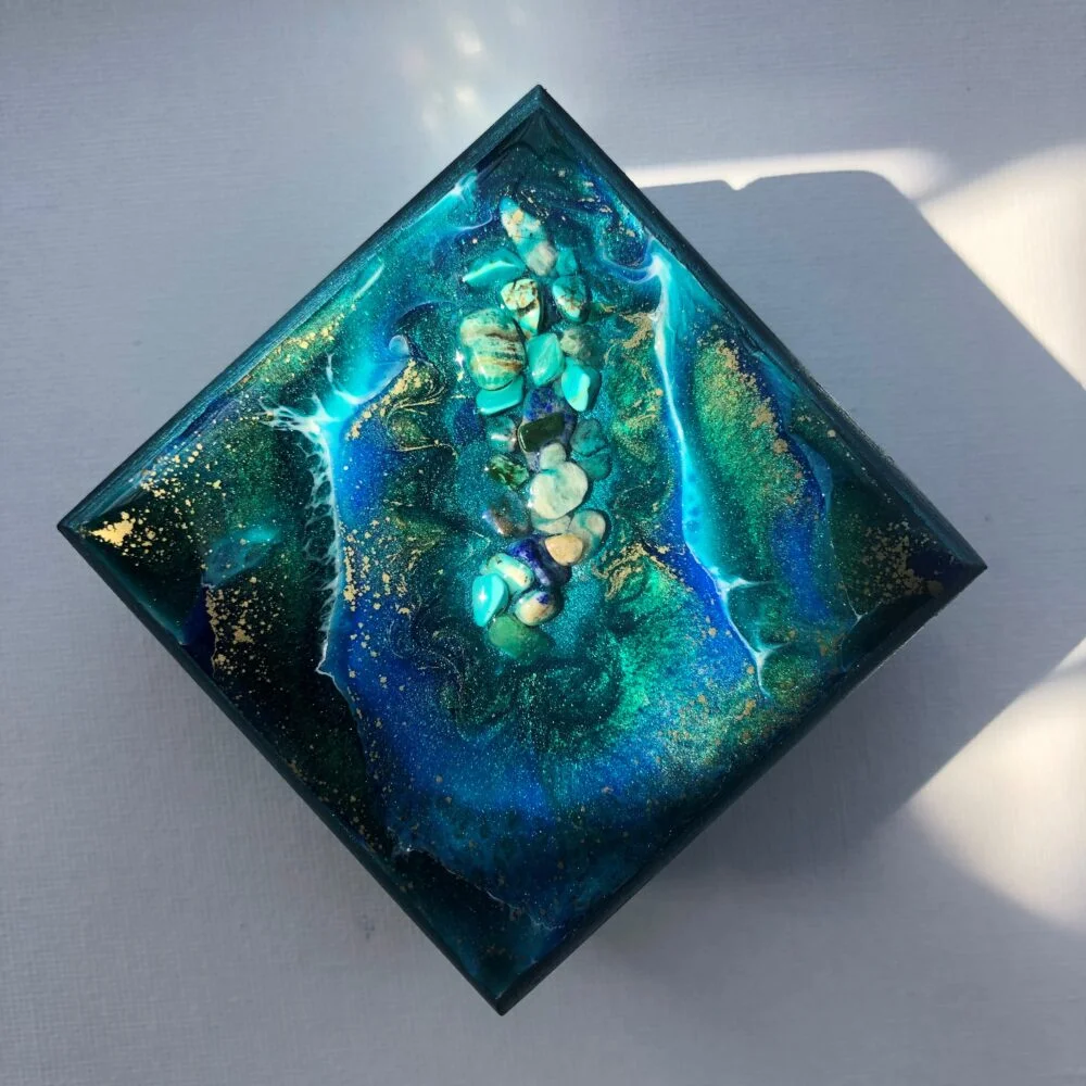 These fabulous trinket boxes are a work of art with each piece having it own unique epoxy resin design. No one piece is ever the same so you can be sure to have a one of a kind piece of artwork which allows you to store small items. This small square hinged wooden box with a magnetic closure, has been hand painted with dark metallic turquoise acrylic paint and wax varnished. The epoxy resin decoration contains natural gemstone shards consisting of Amazonite, Lapis Lazuli and Turquoise. This piece also consists of 3 pigments and gold powder, thus creating a decorative impression of a stoney ocean. The Inside has been lined with waterproof velvet giving a luxurious feel to the inside of this stunning little trinket box. Making it into a wonderful addition to any setting, and an ideal gift for that someone special. Trinket Boxes can also be made to order