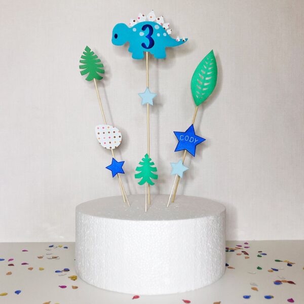 A dinosaur theme cake topper set displayed in a dummy cake. The toppers are personalised with a blue dinosaur with the birthday age and a star with a name. The remaining toppers feature stars and jungle leaves.