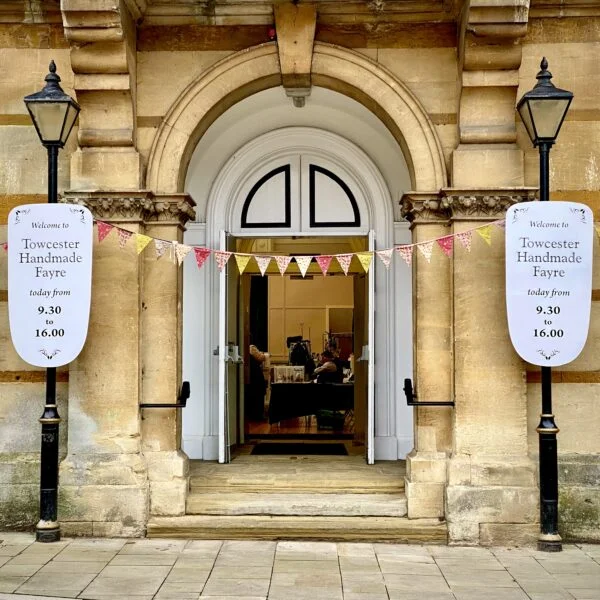 Front of Towcester Town Hall showing Towcester Handmade Fayre banners hung on the old fashioned street lights either side of the main entrance with colourful textile bunting strung between the lights above the steps to the main hall.