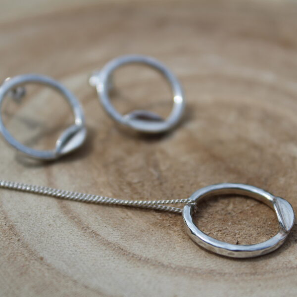 Wood slice with hammered effect hoop necklace with leaf in focus. Out of focus in the background, matching studs.