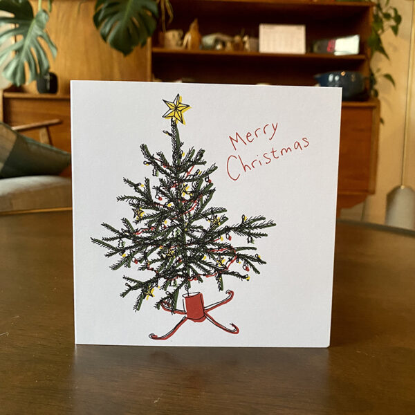 Square white Christmas card stands on a wooden table. Card features a decorated tree illustration
