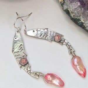 Sterling silver fish earrings, with fish details engraved on the silver, adorned with pink Rhodochrosite Cabochons and Pink keshi Freshwater Pearls. The earrings are placed on a white background with an amethyst stone and spell jar in the background.