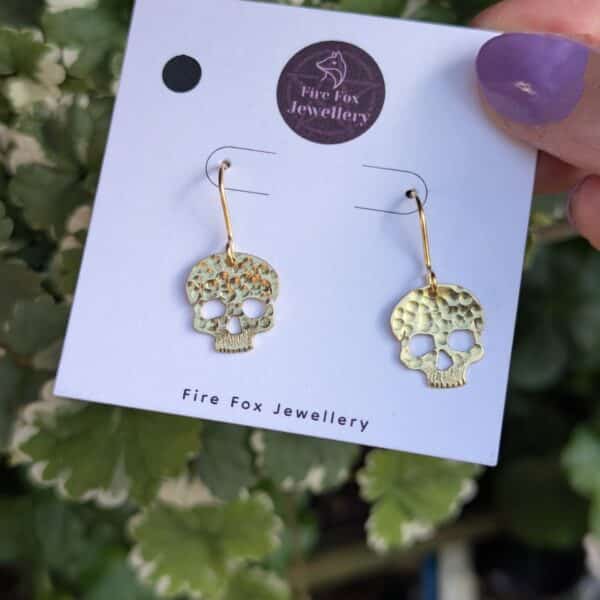 Gold skull earrings with hammered finish on Fire Fox Jewellery card, styled with fresh greenery in the background.