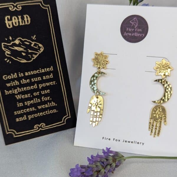 Gold brass earrings placed on a white Fire Fox Jewellery display card to enhance the beautiful detail of each component. Hand-cut from raw gold brass, featuring a star stud, a crescent moon, and a protective hand delicately dangling below. In the background there is purple flowers next to a card that details the symbolism of wearing gold which is success, wealth and protection.