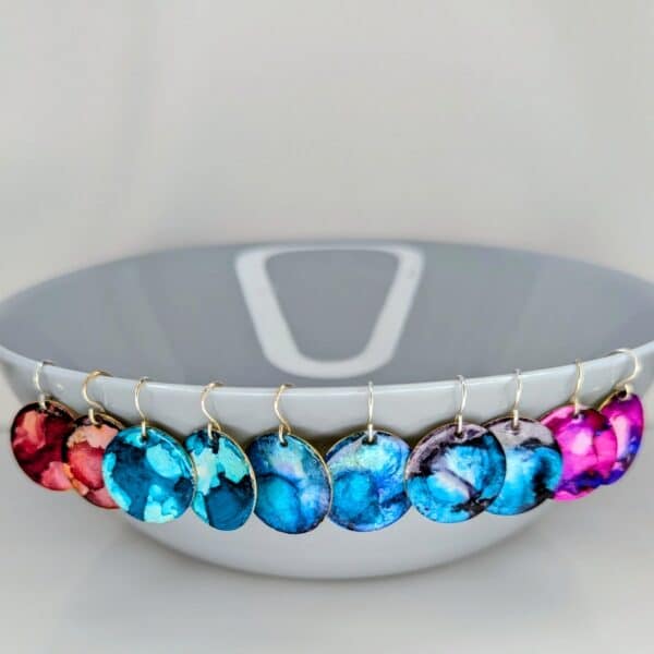 Full collection of colourful enamel earrings, cosmic designs, ocean blues and vibrant pinks/reds with Sterling silver earring hooks.Displayed around a grey bowl