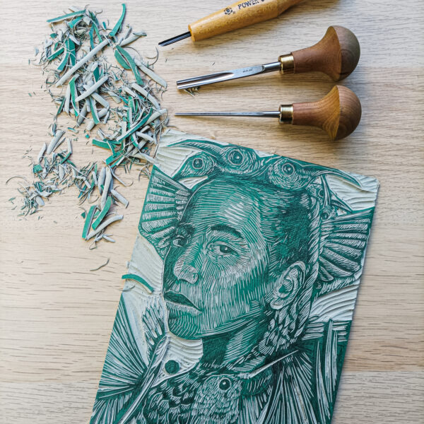 Pice of linocut with a portrait in the process of being carved alongside linocut tools and chippings.