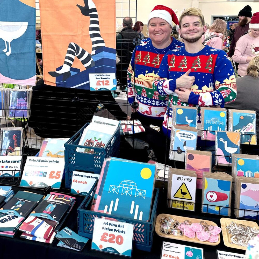 Two independent makers in Christmas jumpers smiling behind a colourful stall of prints, cards, tea towels and gifts at a Shoreham Christmas makers fair.